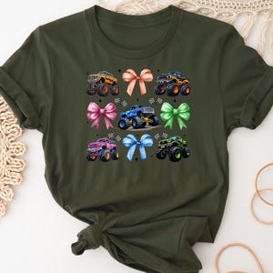 Puede incluir: Camiseta verde oliva con un dise&ntilde;o de monster trucks y lazos. El gr&aacute;fico muestra seis monster trucks de diferentes colores, cada uno con un lazo a juego. Los camiones tienen neum&aacute;ticos grandes y dise&ntilde;os detallados.