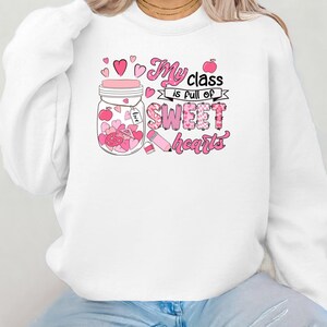 Pu&ograve; includere: Felpa bianca con una grafica rosa di un barattolo pieno di cuori e il testo "My class is full of sweet hearts".