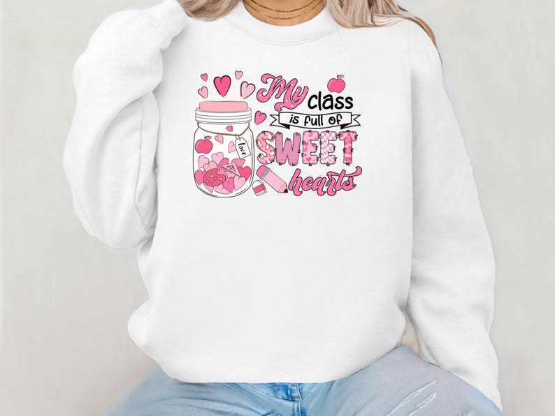 Pu&ograve; includere: Felpa bianca con una grafica rosa di un barattolo pieno di cuori e il testo "My class is full of sweet hearts".