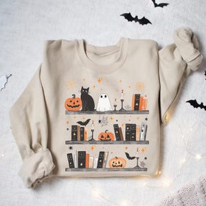 May include: Beige sweatshirt featuring a Halloween-themed bookshelf design. The illustration includes pumpkins, a black cat, a ghost, books, candles, and bats. The design is printed on the front of the sweatshirt.