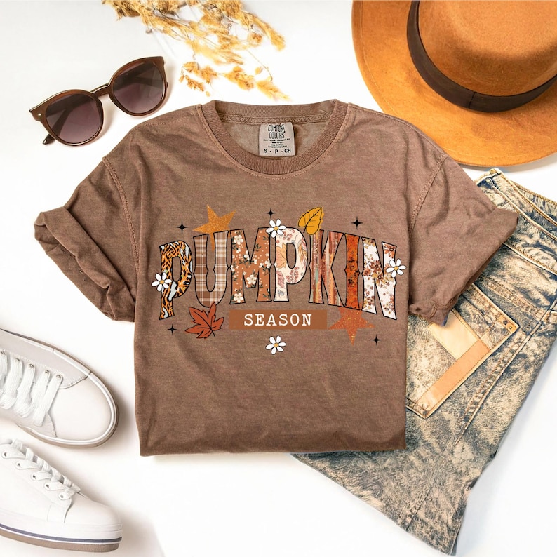 Pu&ograve; includere: Una maglietta marrone con la scritta "PUMPKIN SEASON" in un design a tema autunnale. Il design include motivi floreali, leopardati e a quadri, insieme a stelle e foglie. Occhiali da sole, un cappello, jeans e scarpe da ginnastica sono anche nell'immagine.