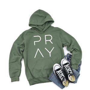 May include: An olive green hoodie with the word "PRAY" in white block letters. The hoodie has a front pocket and a hood. Black canvas sneakers and blue jeans are next to the hoodie on a white background.