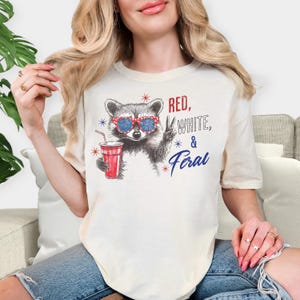 May include: Cream-colored t-shirt featuring a raccoon wearing red, white, and blue sunglasses, holding a red drink, and giving a peace sign. The text "RED, WHITE, & FERAL" is printed on the shirt in red and blue.
