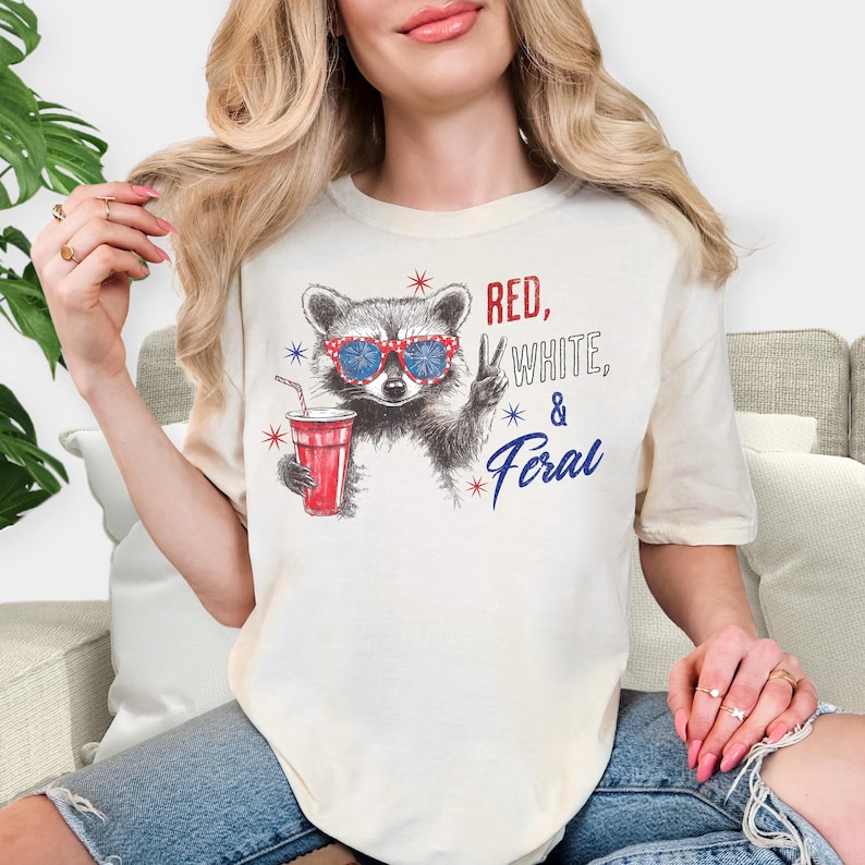 May include: Cream-colored t-shirt featuring a raccoon wearing red, white, and blue sunglasses, holding a red drink, and giving a peace sign. The text "RED, WHITE, & FERAL" is printed on the shirt in red and blue.