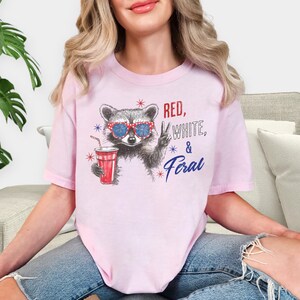 May include: A light pink t-shirt featuring a raccoon wearing sunglasses and holding a red drink. The text "Red, White, & Feral" is printed on the shirt in red, white, and blue.