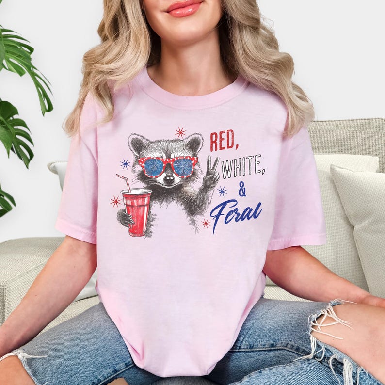 May include: A light pink t-shirt featuring a raccoon wearing sunglasses and holding a red drink. The text "Red, White, & Feral" is printed on the shirt in red, white, and blue.