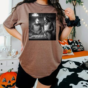 Puede incluir: Camiseta marr&oacute;n con una imagen en blanco y negro de dos figuras con delantales, una de ellas con una m&aacute;scara de hockey. La imagen es una parodia de una escena de pel&iacute;cula de terror. La camiseta la lleva una persona con pantalones cortos negros.