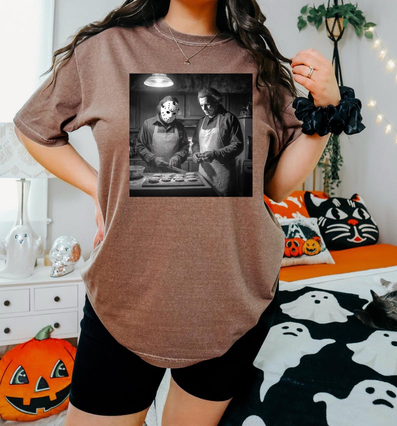 Puede incluir: Camiseta marr&oacute;n con una imagen en blanco y negro de dos figuras con delantales, una de ellas con una m&aacute;scara de hockey. La imagen es una parodia de una escena de pel&iacute;cula de terror. La camiseta la lleva una persona con pantalones cortos negros.