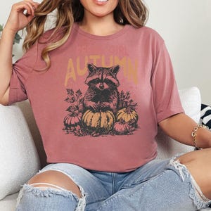 May include: A dusty rose coloured t-shirt featuring a raccoon illustration with pumpkins and the word "AUTUMN". The tee is a casual style, perfect for autumn. The design is vintage.