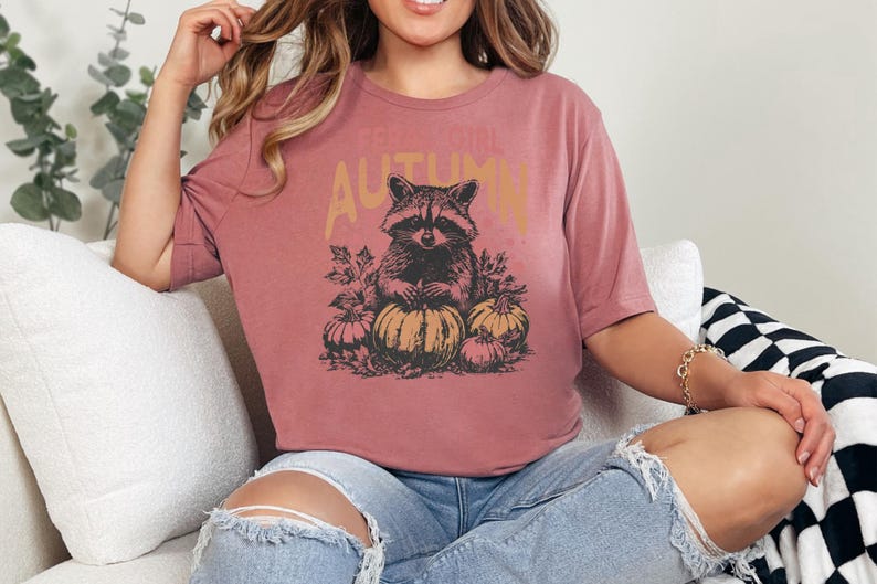 May include: A dusty rose coloured t-shirt featuring a raccoon illustration with pumpkins and the word "AUTUMN". The tee is a casual style, perfect for autumn. The design is vintage.