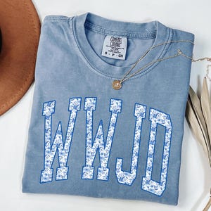 May include: A light blue Comfort Colors t-shirt with the letters WWJD in a white and blue floral pattern. A gold necklace with a pendant is on the shirt. A brown hat is in the upper left corner.