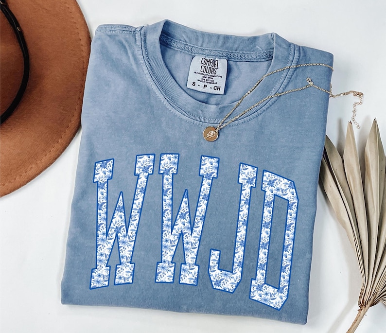 May include: A light blue Comfort Colors t-shirt with the letters WWJD in a white and blue floral pattern. A gold necklace with a pendant is on the shirt. A brown hat is in the upper left corner.