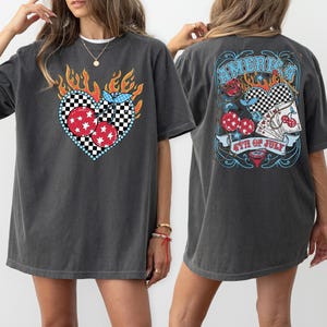 May include: Dark gray t-shirt with a heart-shaped cherry design on the front. The heart is outlined in blue and black checkered pattern, with red cherries and flames. The back has a graphic with the text "America" and "4th of July".