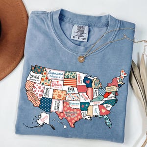May include: A light blue Comfort Colors t-shirt featuring a colorful map of the United States. Each state is decorated with unique patterns and words like "Brave" and "Free". A gold necklace rests on the shirt.