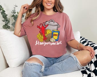 May include: A pink t-shirt with a graphic of a pot of seafood, including a lobster, corn, potatoes, lemon, and garlic. The text "Let the good times boil" is printed below the graphic.