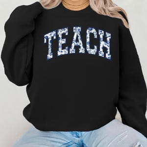 May include: Black sweatshirt with the word "TEACH" in white letters with a blue floral pattern.