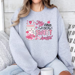 Pu&ograve; includere: Felpa grigia con una grafica rosa di un barattolo pieno di cuori e il testo "La mia classe &egrave; piena di cuori dolci".