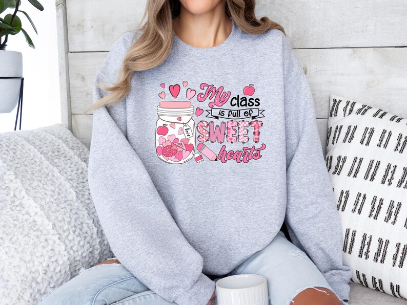 Pu&ograve; includere: Felpa grigia con una grafica rosa di un barattolo pieno di cuori e il testo "La mia classe &egrave; piena di cuori dolci".