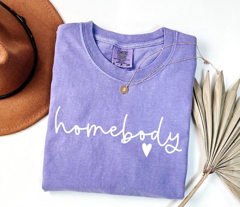 May include: A purple t-shirt with the text "homebody" in white script with a small white heart. The shirt is folded and laid flat on a white surface.