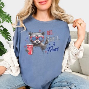 May include: A blue t-shirt features a raccoon wearing sunglasses and holding a red drink. The text on the shirt reads "Red, White, & Feral" in red and blue. The raccoon graphic is in the center of the shirt.