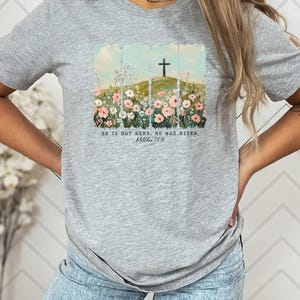May include: A light gray t-shirt featuring a watercolor design of a cross on a hill, surrounded by flowers. The text "HE IS NOT HERE. HE HAS RISEN. Matthew 28:6" is printed below the image. The shirt is made of a soft material.
