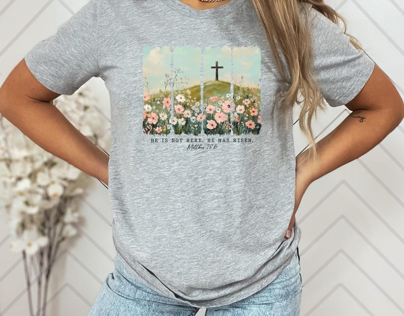 May include: A light gray t-shirt featuring a watercolor design of a cross on a hill, surrounded by flowers. The text "HE IS NOT HERE. HE HAS RISEN. Matthew 28:6" is printed below the image. The shirt is made of a soft material.