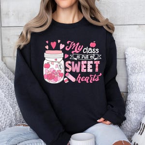Pu&ograve; includere: Felpa nera con una grafica rosa e bianca di un barattolo pieno di cuori e il testo "My class is full of sweet hearts".