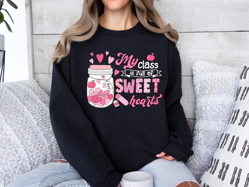 Pu&ograve; includere: Felpa nera con una grafica rosa e bianca di un barattolo pieno di cuori e il testo "My class is full of sweet hearts".