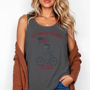 May include: Gray tank top featuring a frog riding a bicycle and holding an American flag. The text "250 YEARS OF FREEDOM 1776-2026" is printed in red. The design is a vintage style.