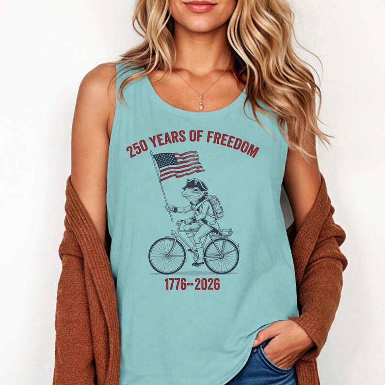 May include: A light blue tank top with a patriotic frog riding a bicycle and holding an American flag. The text "250 YEARS OF FREEDOM" and "1776-2026" are printed on the shirt. The frog is wearing a colonial-style hat and backpack.