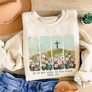 May include: Cream-colored sweatshirt with a floral and cross design. The graphic features a cross on a hill with flowers and the text "HE IS NOT HERE. HE HAS RISEN. Matthew 28:6."