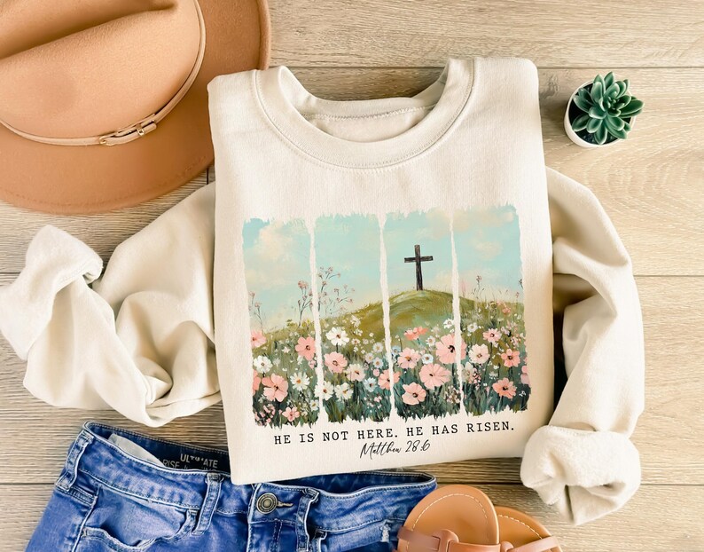 May include: Cream-colored sweatshirt with a floral and cross design. The graphic features a cross on a hill with flowers and the text "HE IS NOT HERE. HE HAS RISEN. Matthew 28:6."