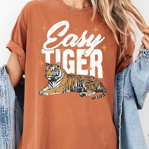 May include: Burnt orange t-shirt with the text "Easy Tiger" in white script above a detailed illustration of a tiger. The tiger is brown and black with a white belly, and is lying down. The shirt has a relaxed fit.