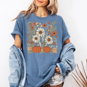 May include: A blue t-shirt featuring a floral design with pumpkins, daisies, and orange flowers. The graphic is centered on the shirt, with a vintage aesthetic. The shirt is made of a soft material.