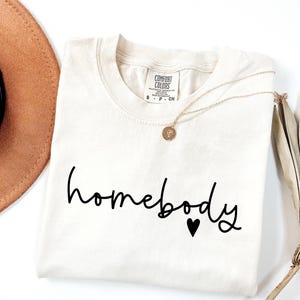 May include: A white t-shirt with the text "homebody" in black script with a small black heart below the text.