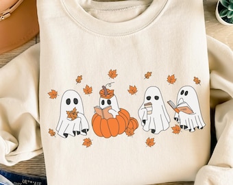 May include: Cream-colored sweatshirt featuring a Halloween-themed design. The graphic includes four white ghosts, orange pumpkins, and fall leaves. One ghost is reading a book, another is holding a leaf, and another is holding a coffee cup.