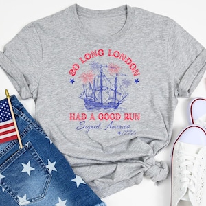 May include: A heather gray t-shirt with the text "SO LONG LONDON HAD A GOOD RUN Signed, America *1776" and a ship graphic. The image also includes denim shorts with white star patterns, a small American flag, and white sneakers.