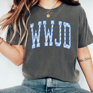 May include: Dark gray t-shirt with the text "WWJD" in a blue floral pattern. The shirt has short sleeves and a crew neck. The letters are in a collegiate style font.