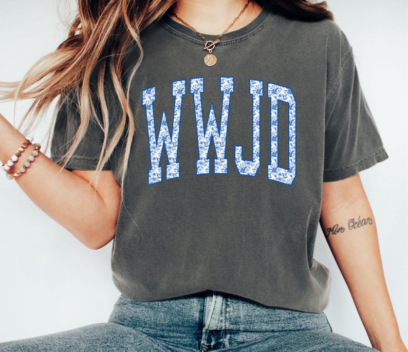 May include: Dark gray t-shirt with the text "WWJD" in a blue floral pattern. The shirt has short sleeves and a crew neck. The letters are in a collegiate style font.