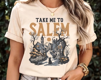 May include: Beige t-shirt with the text "TAKE ME TO SALEM" above a vintage illustration of a spooky town scene. The graphic features houses, trees, a black cat, and flying birds, perfect for Halloween.