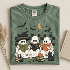 May include: A sage green t-shirt featuring a graphic of three ghosts reading books. The ghosts are surrounded by bats, a crescent moon, and stars. Below them are stacks of books, a candle, a pumpkin, and a black cat. The shirt has a Comfort Colors label.