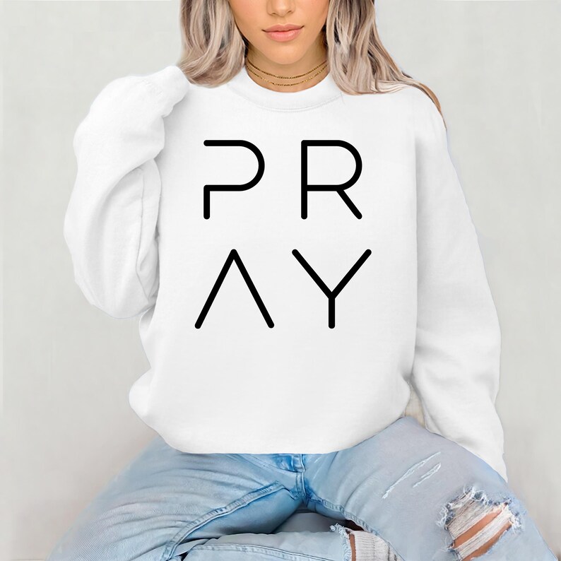 May include: White sweatshirt with the word "PRAY" in black block letters. The letters are arranged in two rows, with "PR" on top and "AY" below. The sweatshirt is a casual style.