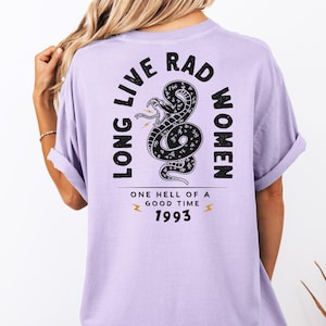 Peut inclure: T-shirt lilas avec un motif noir de serpent et les mots "LONG LIVE RAD WOMEN" et "ONE HELL OF A GOOD TIME 1993". Les manches sont retrouss&eacute;es.