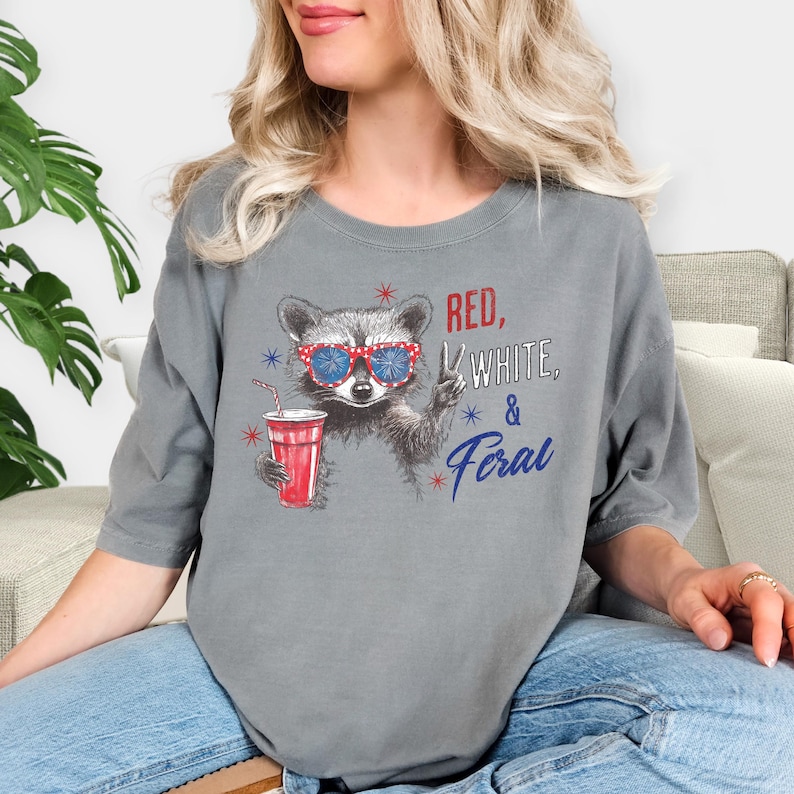 May include: Gray t-shirt featuring a raccoon wearing red, white, and blue sunglasses and holding a red cup. The text "RED, WHITE, & FERAL" is printed on the shirt in red and blue.