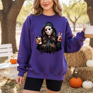 May include: A purple sweatshirt with a graphic of a person in a hockey mask and black hoodie, holding a coffee cup and making a peace sign. The person has long, dark hair. Pumpkins and hay bales are in the background.