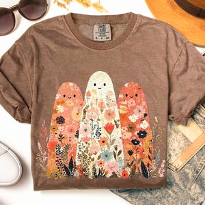 May include: Brown t-shirt featuring a whimsical design of three ghost-like figures adorned with floral patterns in shades of pink, orange, and white. The shirt has a crew neck and short sleeves. The tag reads "COMFORT COLORS".