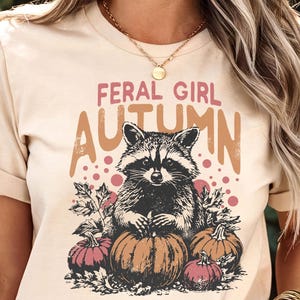May include: Beige t-shirt featuring a raccoon illustration with the text "FERAL GIRL AUTUMN". The design includes pumpkins and autumn leaves. The top is a short-sleeved crew neck.