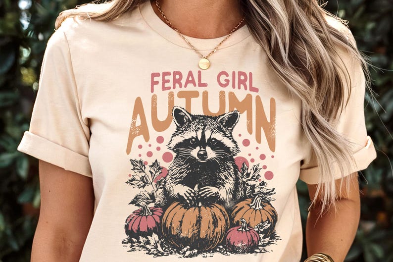 May include: Beige t-shirt featuring a raccoon illustration with the text "FERAL GIRL AUTUMN". The design includes pumpkins and autumn leaves. The top is a short-sleeved crew neck.