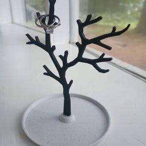 May include: A black metal tree-shaped ring holder with a white speckled base. Two rings are displayed on the branches.