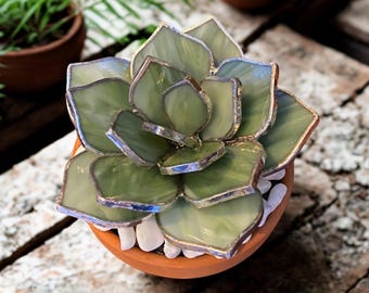 May include: A handcrafted glass succulent sculpture in a terracotta pot. The succulent features light green, textured glass petals with silver-colored edging. White pebbles surround the base of the succulent. The pot is sitting on a wooden surface.